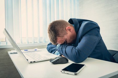 Un hombre de negocios cansado durmiendo en su oficina. Un hombre con chaqueta azul duerme por la mañana en el lugar de trabajo. El empleado se durmió en la mesa delante del portátil