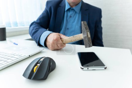 Photo for A businessman sitting in a table with a hammer in his hand ready to crash it on a smartphone - Royalty Free Image