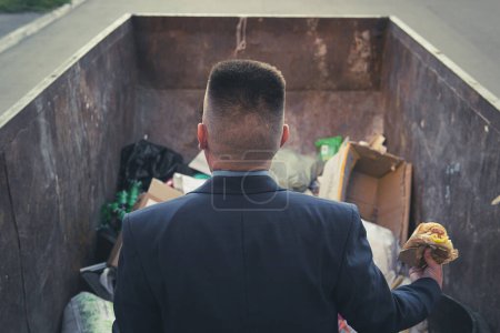 Photo for An impoverished businessman rummages in a trash can in search of food. A homeless homeless man in a suit holds a hamburger in his hand while standing in a landfill. bankruptcy, no money concept - Royalty Free Image