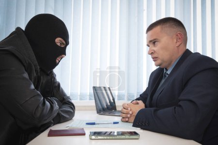 Photo for A businessman and robbers are sitting at a table. A racketeer in a black balaclava forces to sign a contract. The concept of a raider takeover of the company. signing of important documents. - Royalty Free Image