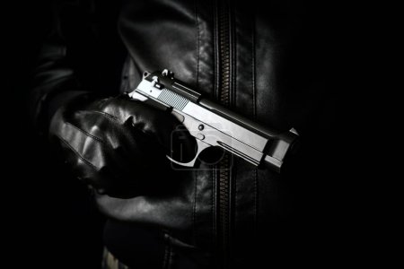 Photo for The concept of the crime of banditry. A dangerous shooter and a black pistol on a dark background. The hired killer is preparing to shoot. Pulls a firearm out of his jacket pocket - Royalty Free Image