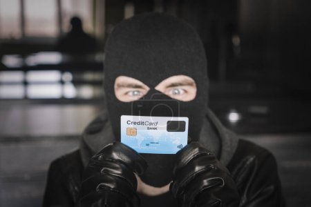 Foto de Delincuente enmascarado calificado usando una tarjeta de crédito robada para comprar cosas en línea. Un ladrón con una máscara negra tiene una tarjeta robada en sus manos. - Imagen libre de derechos