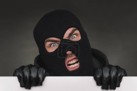 Photo for An evil thief in a mask looks out from behind a white board. an evil bandit with a mask on his face. - Royalty Free Image