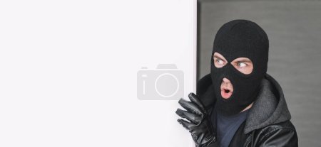 Photo for An evil thief in a mask looks out from behind a white board. Space for text and design. copy space. the surprised bandit looks away - Royalty Free Image
