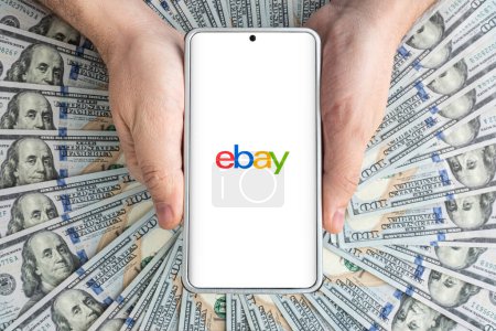 Foto de El logotipo de la página web de eBay en la pantalla del teléfono inteligente en las manos de los hombres que yacen en dólares estadounidenses. 30 de marzo de 2023. Barnaul. Rusia. - Imagen libre de derechos