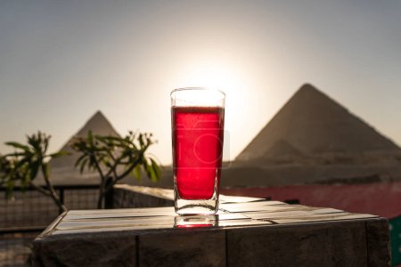 Foto de Carkade tradicional de té de hierbas árabe en un vaso sobre el fondo de las grandes pirámides de Giza. Hermosa vista de la atracción en el restaurante al atardecer. El sol se pone detrás de las pirámides - Imagen libre de derechos