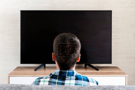 Téléchargez les photos : Vue de dos de l'homme regardant la télévision. Plasma à écran plat avec écran vide. Assis sur le canapé à la maison. Homme méconnaissable regardant un film le week-end. Focus sélectif - en image libre de droit