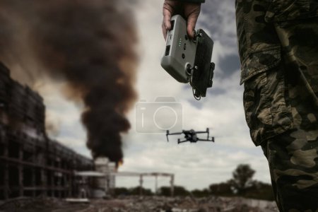 Foto de El operador de UAV con un mando a distancia está parado contra el fondo de un edificio destruido con humo negro en la ciudad. El concepto de tecnologías no tripuladas en la guerra moderna. bombardeo dron de reconocimiento - Imagen libre de derechos