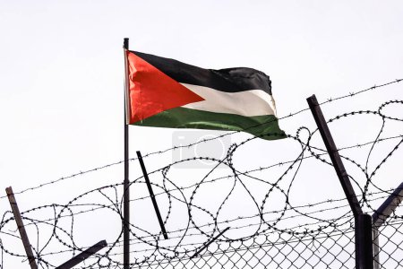 Foto de Vista de la bandera palestina detrás del alambre de púas contra el cielo nublado. puesto fronterizo en la frontera de Palestina. El puesto de control de la embajada del país. la frontera palestino-israelí. - Imagen libre de derechos