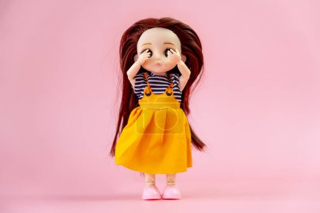 Photo for A child toy doll with dark hair in an orange dress standing on a pink background. Plastic children's toy. Doll games for imagination. the frightened doll closes her eyes in fear. - Royalty Free Image