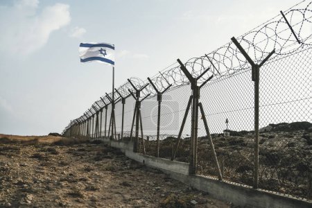 View of israeli flag behind barbed wire in the desert against cloudy sky.