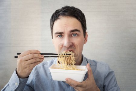 Photo for A young man in a blue shirt is eating noodles from a box with a dissatisfied face. Lunch at the office. tasteless junk food - Royalty Free Image