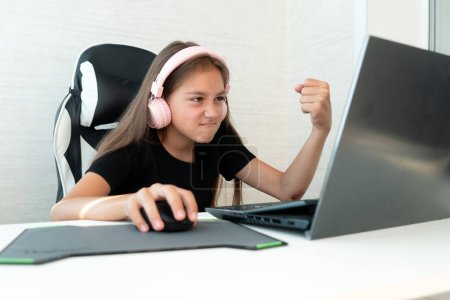 Photo for A teenage girl in pink headphones is playing computer games on a white background. A negative emotion on the face. the child gamer shows his fist at the laptop screen. - Royalty Free Image