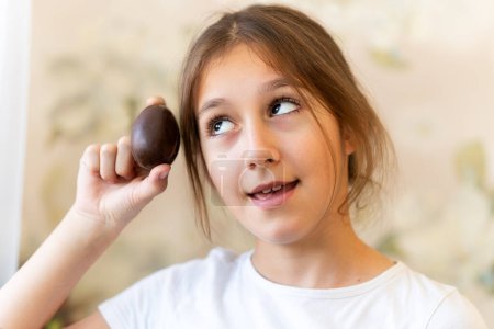 Foto de Un niño abre un huevo de chocolate. La chica está sorprendida por la sorpresa. La niña sostiene un huevo de chocolate en sus manos y espera una buena sorpresa - Imagen libre de derechos