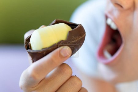 Téléchargez les photos : Une petite fille mord un œuf en chocolat avec un jouet en plastique à l'intérieur. aliments dangereux dangereux pour les enfants - en image libre de droit