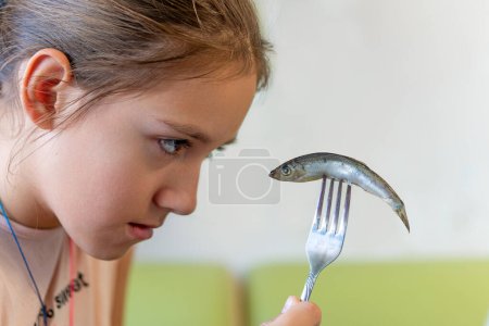 Fiese Fische. Kinder essen nicht gern Fisch. Ein junges Mädchen blickt mit Verachtung auf eine Sprotte auf einer Gabel in ihrer Hand.