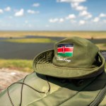 The national flag of Kenya on a green hat and a backpack lying on a table against a beautiful African landscape. The concept of safari and adventure in the Kenyan Republic.