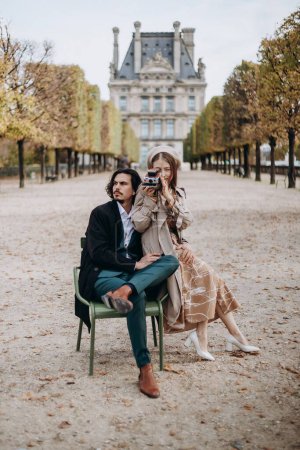 Téléchargez les photos : Un couple amoureux se promène dans les rues de Paris, un mec en manteau et costume, une fille en robe avec un béret et un trench coat en automne avec caméra - en image libre de droit