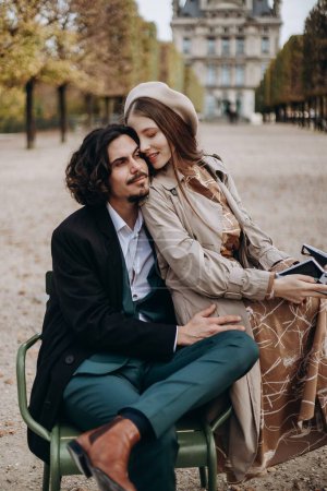 Téléchargez les photos : Un couple amoureux se promène dans les rues de Paris, un mec en manteau et costume, une fille en robe avec un béret et un trench coat en automne avec caméra - en image libre de droit