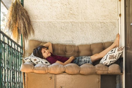 Photo for Barefoot Kid Having an Afternoon Rest on the Couch on the Outdoor Patio. Having Lazy Summer in the Cottage in Counryside. Copy Space on the top - Royalty Free Image