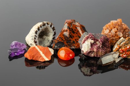 Photo rapprochée du groupe de pierres et minéraux, y compris Jaspe rouge, Aragonite Spoutnik, Cristaux d'érythrite, Obsidienne d'acajou et Géode d'agate sur la surface réfléchissante