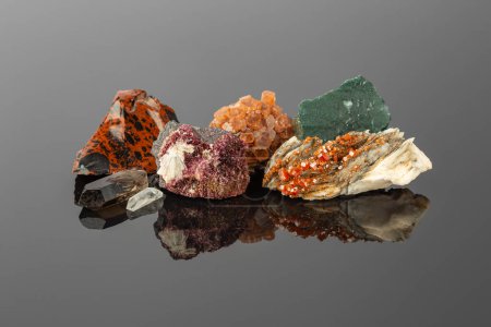 Groupe de pierres et de minéraux disposés en rangée, y compris la vanadinite sur barytine, l'aragonite spoutnik, les cristaux d'érythrite, l'acajou obsidienne, le quartz fumé et l'agate de mousse sur la surface réfléchissante
