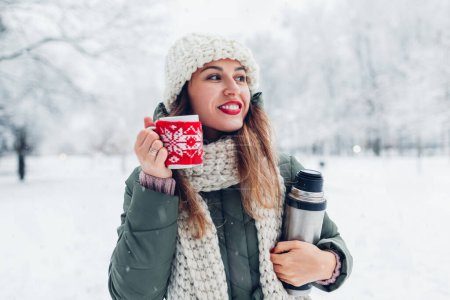 Foto de Retrato de una joven mujer feliz bebiendo té caliente sosteniendo frasco de vacío en el parque de invierno nevado disfrutando del paisaje bajo la nieve que cae. Copa vestida en rojo caso de Navidad de punto - Imagen libre de derechos