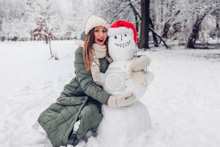 Photo for Happy young woman hugs snowman in snowy winter park dressed in Christmas Santa hat. Festive fun outdoor activities during cold weather - Royalty Free Image
