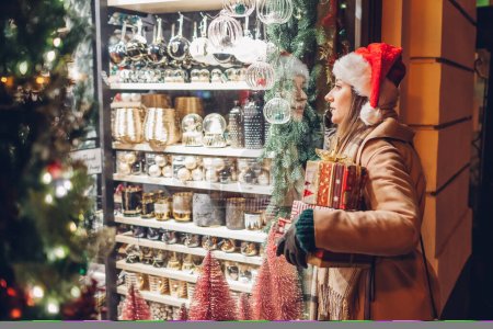Téléchargez les photos : Femme curieuse dans le chapeau du Père Noël tenant cadeaux boîtes-cadeaux regardant vitrine de magasin sur la rue de la ville à la saison de Noël. Fille shopping pour le Nouvel An décor de vacances - en image libre de droit