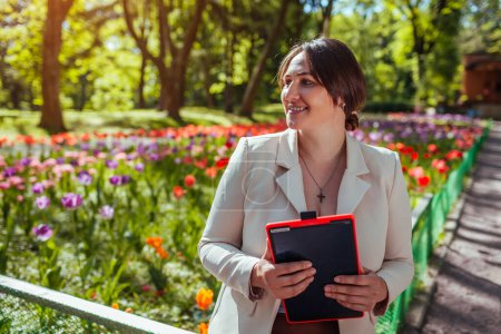 Portrait of happy businesswoman holding tablet sitting in spring park. Female landscape designer enjoys results of blooming flowerbeds in public garden