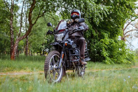 Photo for Biker unfastens helmet after riding motobike in summer forest. Man wearing leather clothes outdoors. Rider sitting on transport. Summer road trip - Royalty Free Image
