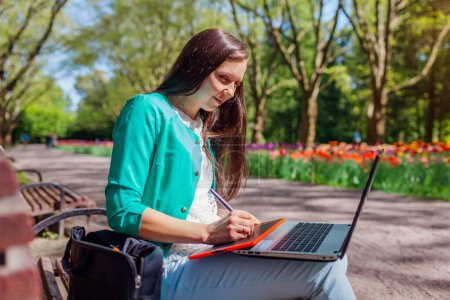 Outdoor portrait of student studying in spring park using laptop and tablet. Young woman getting ready for lecture in public garden