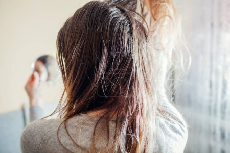 Frau überprüft schmutzige ölige und fettige Haare und schaut zu Hause in den Spiegel. Kosmetikkonzept für schlechte Haarpflege. Rückseite
