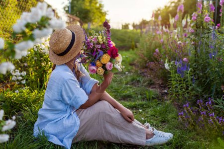Woman gardener enjoying summer garden at sunset holding fresh roses mixed with veronika, foxgloves. Farmer picked bouquet of flowers and relaxing on grass path