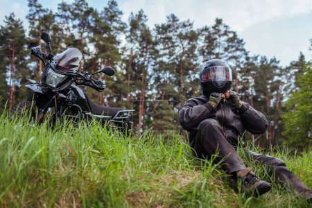 Photo for Close up of biker unfastening helmet after riding motobike in summer forest. Man wearing leather clothes outdoors. Safety measures - Royalty Free Image