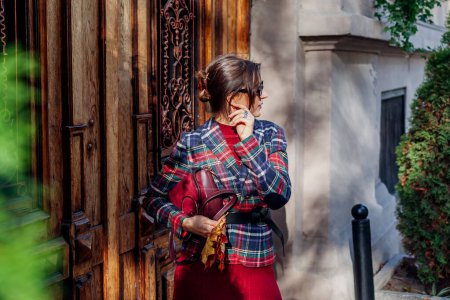 Portrait of stylish woman wearing hair claw clip holding red purse wearing checkered blazer, accessories on autumn city street. Fall fashionable old money outfit