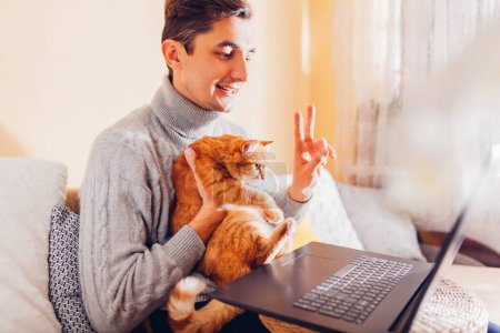 Homme facetiming de la maison tenant animal de compagnie à l'aide d'un ordinateur. Chat roux regardant l'écran de l'ordinateur portable pendant le chat vidéo. Communication en ligne.
