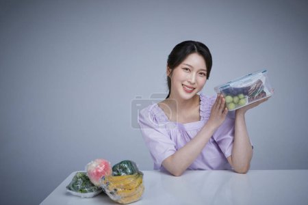 koreanisch asain schöne Frau hält eine Plastikwanne mit Obst und Gemüse in Saranfolie gewickelt.