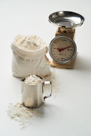 A small bag and measuring cup are filled with flour in front of the scale