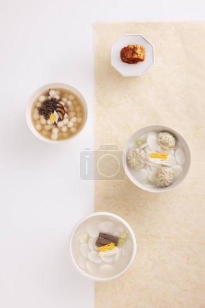 On a white table, three bowls of rice cake soup are placed with kimchi