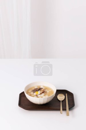 In a wooden tray on a white table, there are rice cake soup and spoons