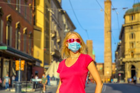 Téléchargez les photos : Lifestyle femme avec masque chirurgical dans Via Ugo Bassi route avec deux tours sur fond. COVID-19 Coronavirus après quarantaine avec éloignement social dans la ville italienne de Bologne
. - en image libre de droit