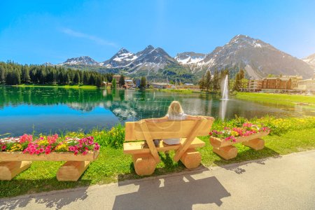 Woman tourist on a park bench of Arosa town at dawn. Touristic resort by Obersee Lake in Switzerland. Arosa lakefront by cable car station to Aroser Weisshorn peak. Plessur Region in Grisons Canton.