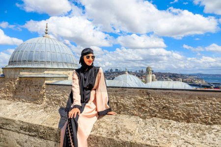 Sitting on a stone wall at Suleymaniye Mosque, a woman in an Arab hijab outfit enjoys the panoramic view of Istanbul. Tranquility and harmony of the historic UNESCO landmark.