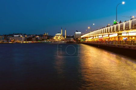 Evening view of galata bridge with glowing lights and the istanbul silhouette against the twilight sky wih Yeni Cami new mosque.