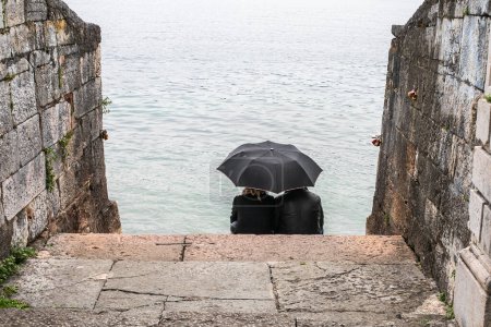 Un par de jóvenes se sientan bajo un paraguas negro en la orilla del lago en un día lluvioso, vista trasera. Espié el marco. Foto de alta calidad