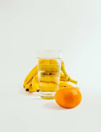 Foto de Plátanos, naranja y lujoso vaso de agua sobre fondo blanco. Composición vertical mínima, concepto de estilo de vida saludable desintoxicación - Imagen libre de derechos
