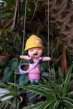 Téléchargez les photos : Jolie poupée en terre cuite accrochée à un arbre. Poupée enfant en faïence colorée sur swing.home décoration de jardin et sculpture dans le concept de jardin - en image libre de droit