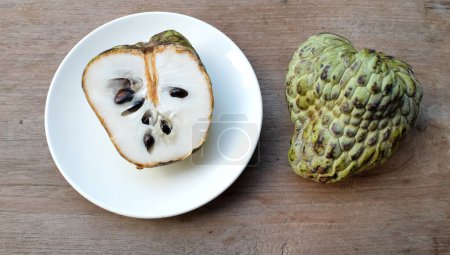 fresh green raw custard apple,sugar apple, annona cherimola (annona squamosa l.) fruit cut in half sliced with leaves in a white plate on wooden table backdrop. tropical exotic fruit and health
