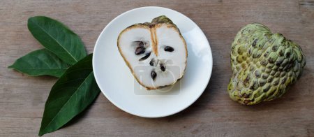 fresh green raw custard apple,sugar apple, annona cherimola (annona squamosa l.) fruit cut in half sliced with leaves in a white plate on wooden table backdrop. tropical exotic fruit and health